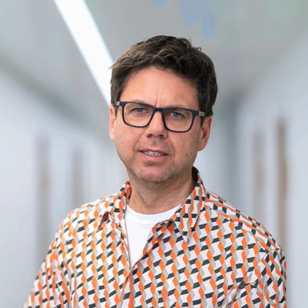 Ein Mann mit Brille und buntem Hemd steht in einem hellen, verschwommenen Flur.