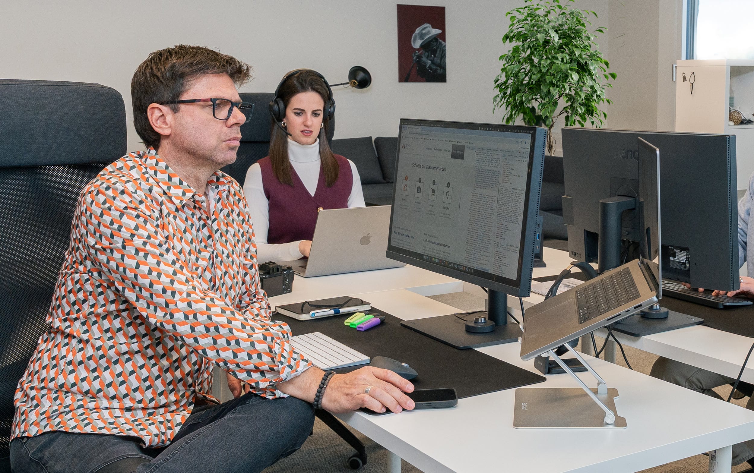 Zwei Personen arbeiten an Computern in einem modernen Büro, auf deren Schreibtischen sich Büromaterial befindet.